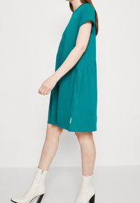 Tyrkysové šaty s krátkými rukávy, volným střihem, nařaseným pasem a jemnými bočními kapsami. Nosené s bílými kotníkovými botami a hladkou texturou.