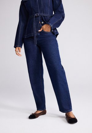 Person iført en mørkeblå denimjakke med bælte, matchende højtaljede straight-leg jeans og sorte balletsko med brune kanter.