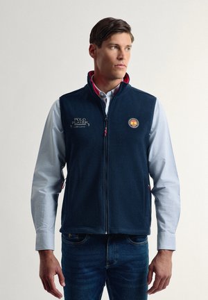 Chaleco de forro polar marino con cuello alto, cremallera frontal y bolsillos laterales; presenta logotipos bordados en acentos dorados y rojos.