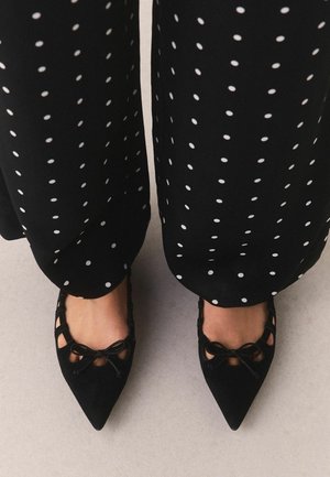 Pieds portant des ballerines noires pointues avec un détail nœud, associées à un pantalon noir à pois blancs.