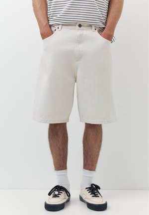 Mężczyzna w kremowych szortach do kolan, białych skarpetkach i białych trampkach z czarnymi podeszwami i sznurówkami, ręce w kieszeniach, na wysokości talii widoczna koszulka w paski.