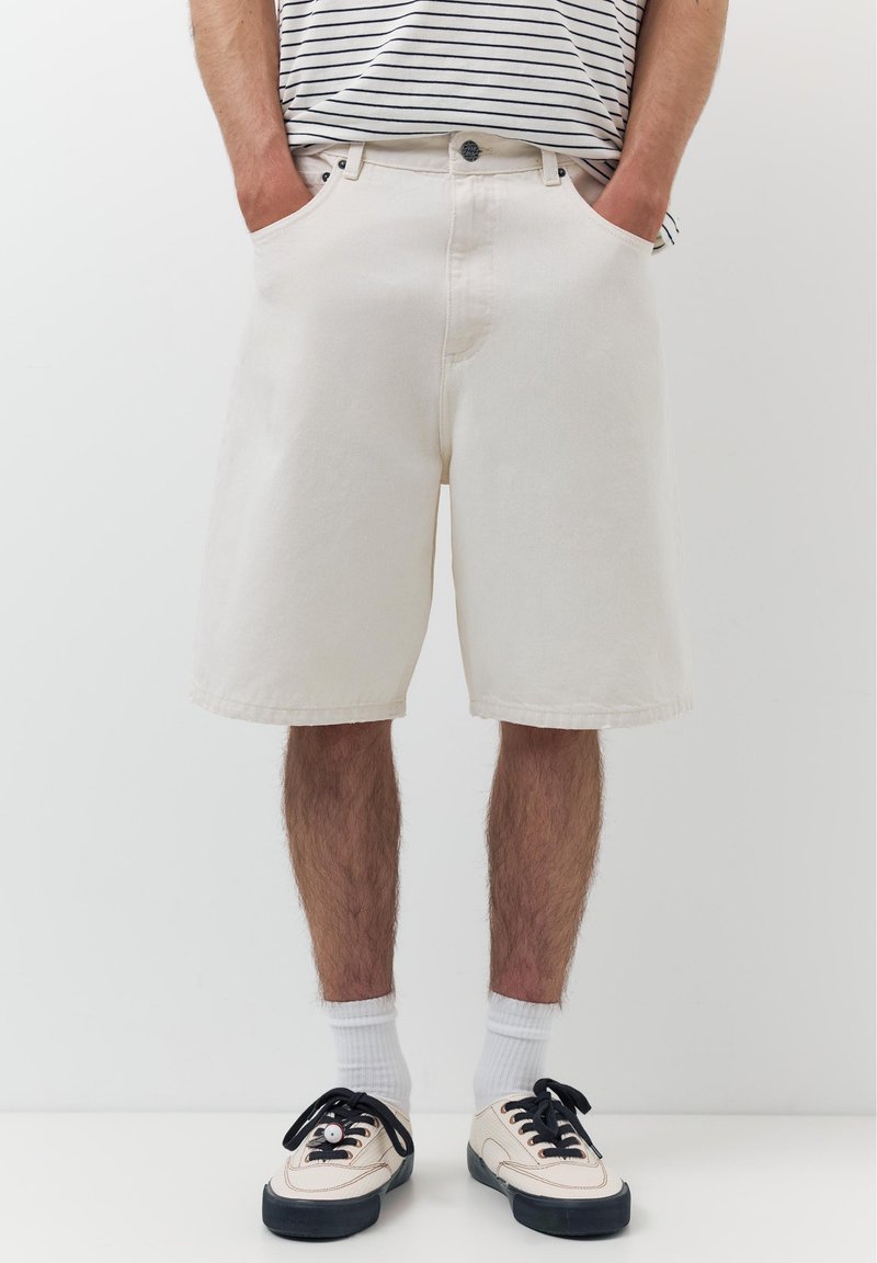 Homme portant un short blanc cassé jusqu'aux genoux, des chaussettes blanches et des baskets blanches avec des semelles et des lacets noirs, les mains dans les poches, chemise rayée visible à la taille.
