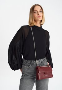Blusa nera con maniche trasparenti, jeans grigi a vita alta e una piccola borsa in pelle bordeaux con tracolla a catena e chiusura a pattina.