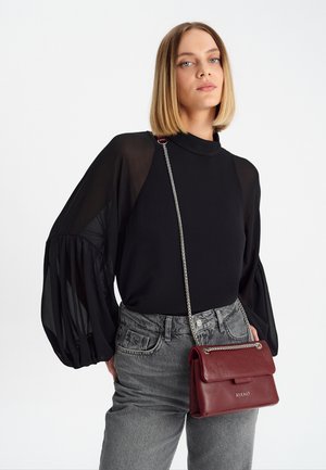 Schwarze Bluse mit durchsichtigen Ärmeln, graue hochgeschnittene Jeans und eine kleine bordeauxfarbene lederne Handtasche mit Kettenriemen und Klappenverschluss.