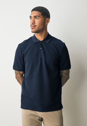 Muž s tetovaniami nosí tmavomodré polo tričko a béžové nohavice, pozerá sa doľava na jednoduchom svetlom pozadí.