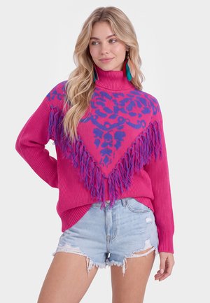 Ung kvinde med langt blondt hår bærer en knaldpink turtleneck sweater med lilla mønstre og frynser, kombineret med lyseblå slidte denimshorts.