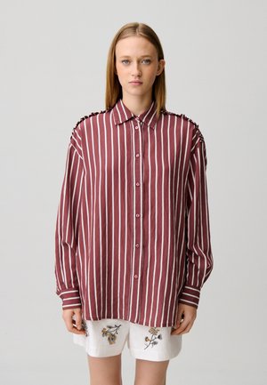 Femme portant une chemise ample couleur bordeaux avec des rayures blanches verticales et un short blanc orné de broderies florales, se tenant devant un fond uni.