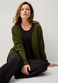 Cardigan verde oliva realizzato in materiale morbido e testurizzato, con design aperto, sovrapposto a una maglietta nera, abbinato a pantaloni neri.