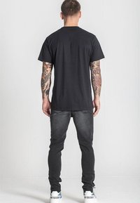 Čierne bavlnené tričko s okrúhlym výstrihom, v kombinácii s tmavošedými džínami a bielymi teniskami s modrými akcentmi. Viditeľné tetovania na ramenách.