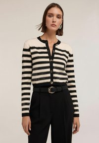 Randig kofta i krämvitt och svart, med knäppning framtill och ribbad textur. Kombinerad med högmidjade svarta byxor och ett bälte.