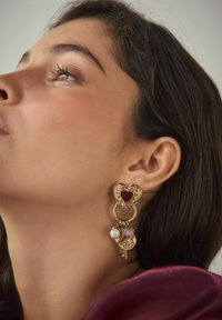 Goldfarbene Ohrringe mit einem herzförmigen roten Mittelpunkt, dekorativem Metallwerk und hängenden Elementen mit pinken und weißen Perlen.