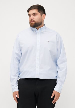 Camisa de botones azul claro con estampado pequeño, hecha de algodón, que cuenta con un cuello clásico y un logo en el pecho; combinada con pantalones negros.