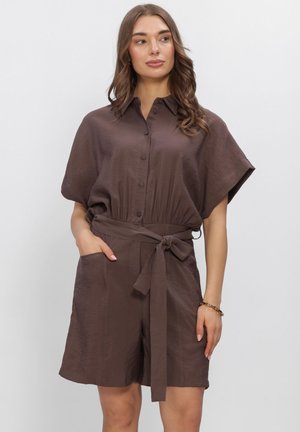 Femme portant une combinaison-short marron à manches courtes ceinturée avec des boutons sur le devant, debout avec une main dans la poche sur fond blanc.