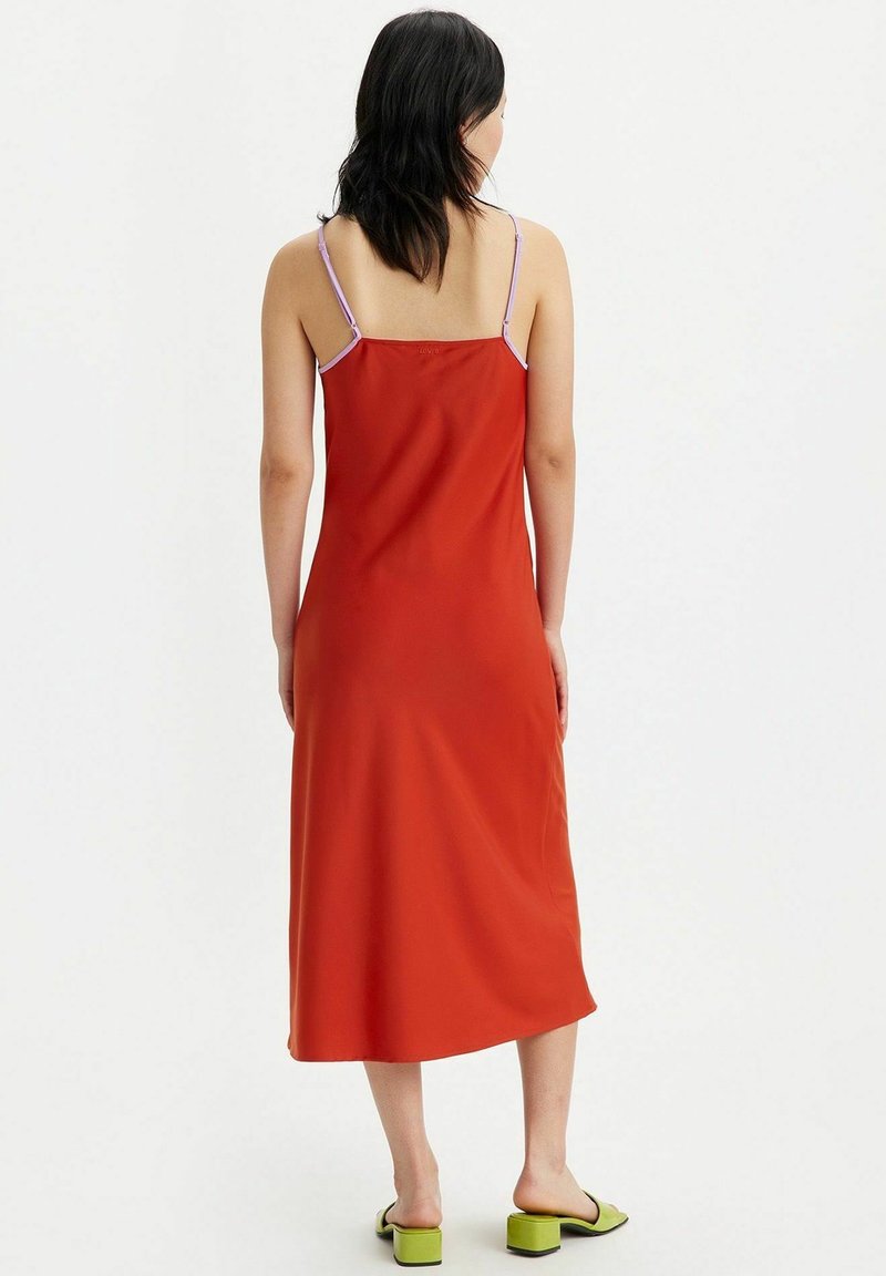 Femme aux cheveux noirs mi-longs portant une robe midi rouge sans manches avec de fines bretelles lavande et des sandales vertes à talons blocs, de dos.