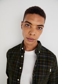 Green and navy plaid shirt with buttoned collar, paired with a white crew neck t-shirt. Features a chest pocket and textured fabric.