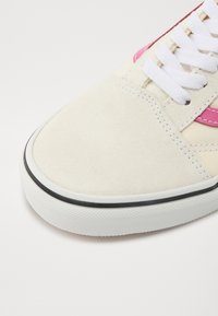Beige ruskindsneaker med hvide snørebånd, lyserøde accenter og en sort gummisål. Har en struktureret tåhætte og syede detaljer.