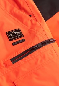 Oranje waterdichte jasstof met zwarte rits, voorzien van een zwart logo-opdruk met bergontwerp en zwarte accenten op de borst.