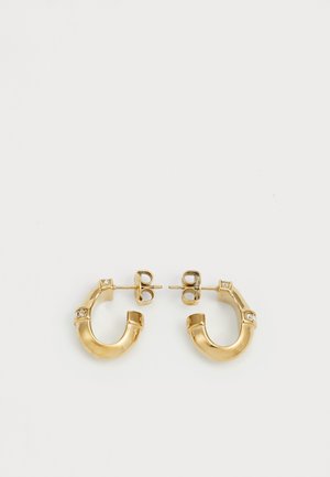 Paire de petites boucles d'oreilles créoles en or avec deux pierres précieuses claires incrustées sur chaque créole, fermées par des fermoirs à poussette sur un fond blanc.