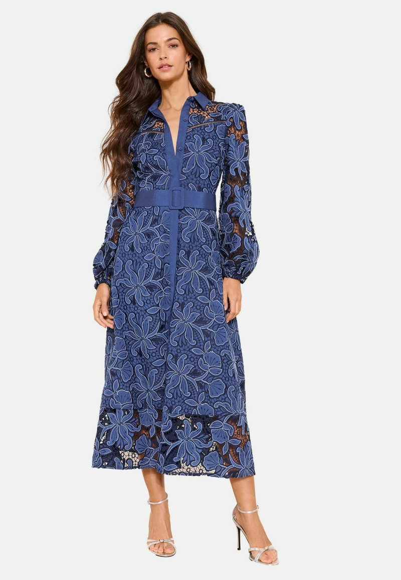 Femme portant une robe en dentelle bleu marine à manches longues avec broderie florale, une ceinture assortie et des sandales à talons hauts argentées, debout avec une expression neutre.