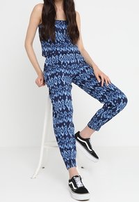 Niebieski, wzorzysty dwuczęściowy zestaw, składający się z topu bez ramiączek oraz spodni w stylu jogger, oba z geometrycznym wzorem. Uzupełniony czarnymi sneakersami.