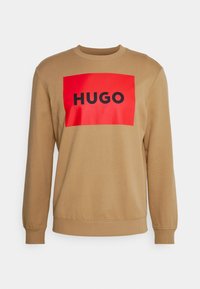 Sudadera beige con cuello redondo, mangas largas y un gran logo rectangular rojo que muestra la palabra "HUGO" en letras negras.