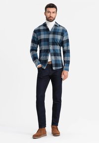 Camisa de cuadros azul y gris, de botones y de mangas largas, llevada sobre un cuello de tortuga blanco. Jeans oscuros y botas de ante marrón completan el conjunto.