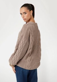 Beige kabelgebreid sweater met textuurpatronen en een ronde halslijn. Losse pasvorm, lange mouwen en geribbelde manchetten. G gedragen met donkerblauwe jeans.