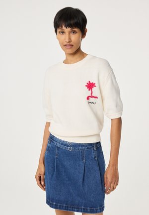 Frau mit kurzen schwarzen Haaren, die einen weißen Pullover mit rosa Blumenstickerei und einen blauen Jeansrock mit herzförmigen Gürteldetails trägt.