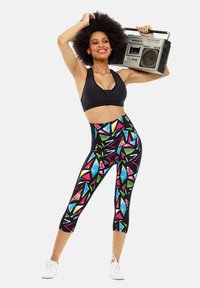 Sujetador deportivo negro y leggings de cintura alta con un patrón geométrico multicolor. La modelo sostiene una radiocassete y lleva zapatillas blancas.