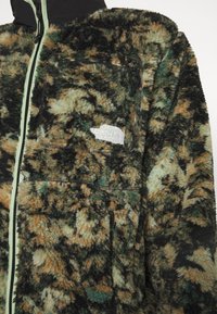 Fleecejacke im Tarnmuster in Orange, Grün und Schwarz. Verfügt über eine vordere Zipp-Tasche und einen schwarzen Kragen. Markenlogo auf der Brust.