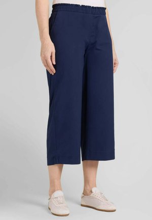 Persona che indossa pantaloni navy blue ampi e corti con elastico in vita e sneakers beige chiaro su sfondo grigio uniforme.