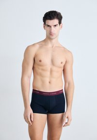 Schwarze Baumwoll-Boxershorts mit burgunderrotem elastischem Bund, der das Logo "TOMMY HILFIGER" trägt. Enganliegendes Design und glatte Textur.