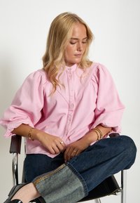 Růžová košile na knoflíky s nařasenými rukávy, vyrobená z texturované látky. Nosená s džíny, v doplněné zlatými doplňky a uvolněným střihem.