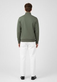 Grön, zip-up sweatshirt med ståkrage, åtsittande ärmar och fåll. Parat med vita byxor och svarta skor, sedd bakifrån.