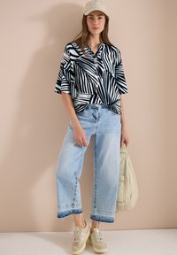 Femme debout portant une chemise à manches courtes à motif noir et blanc, un jean large bleu clair, des baskets beige, une casquette et tenant un sac beige.