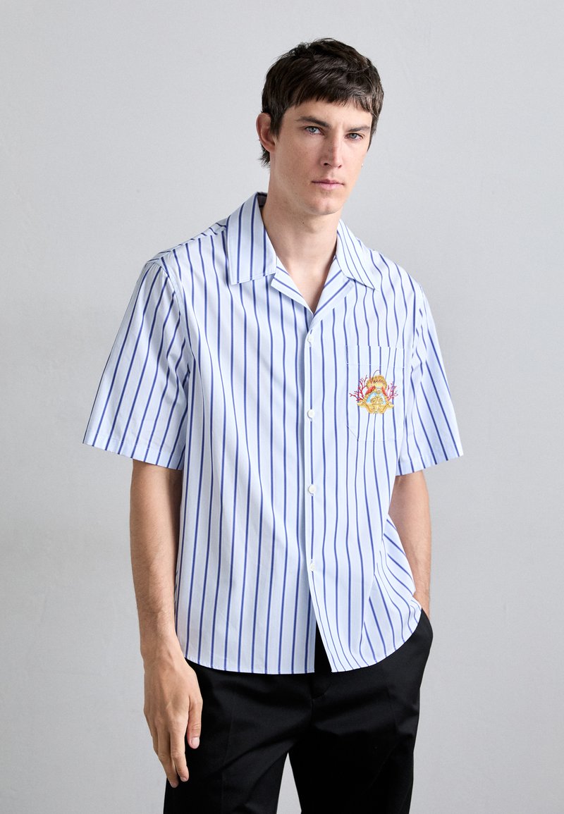 Short-sleeve button-up shirt in light blue with vertical white stripes. Features an embroidered design on the left chest pocket.