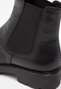 Bottine en cuir noir avec un panneau élastique noir texturé, une semelle en caoutchouc robuste et des coutures détaillées le long du bord supérieur.