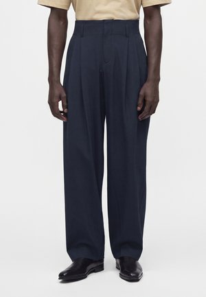 Homme portant un pantalon bleu marine taille haute et coupe ample, associé à des chaussures habillées noires et une chemise beige à manches courtes, debout devant un fond uni.