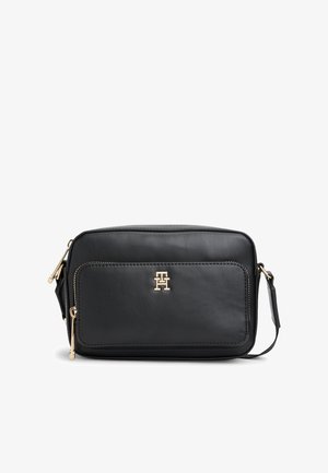 Melna taisnstūra ķermeņa soma ar priekšējo rāvējslēdzēja kabatu, zelta logotipa detaļu un regulējamu plecu siksnu.