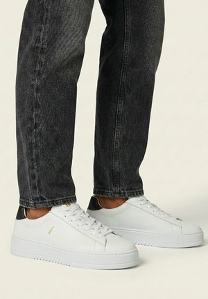 Witte lage sneakers met zwarte hielaccenten gedragen bij zwarte jeans op een lichte achtergrond, met zij- en schuine vooraanzichten.