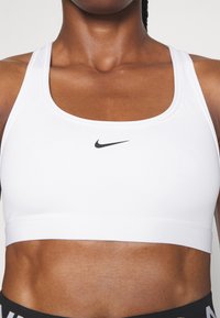 White sports bra made of stretchy fabric with a smooth texture. Features a curved neckline and a black Nike logo on the front.
