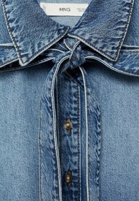 Chemise en denim à col noué de couleur bleu clair. Présente des coutures visibles, une fermeture boutonnée sur le devant et un motif de tissu texturé.