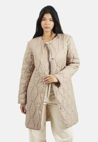 Giacca lunga trapuntata beige con colletto rotondo, chiusure a pressione e motivo ondulato orizzontale. Indossata sopra una maglietta bianca e pantaloni chiari.