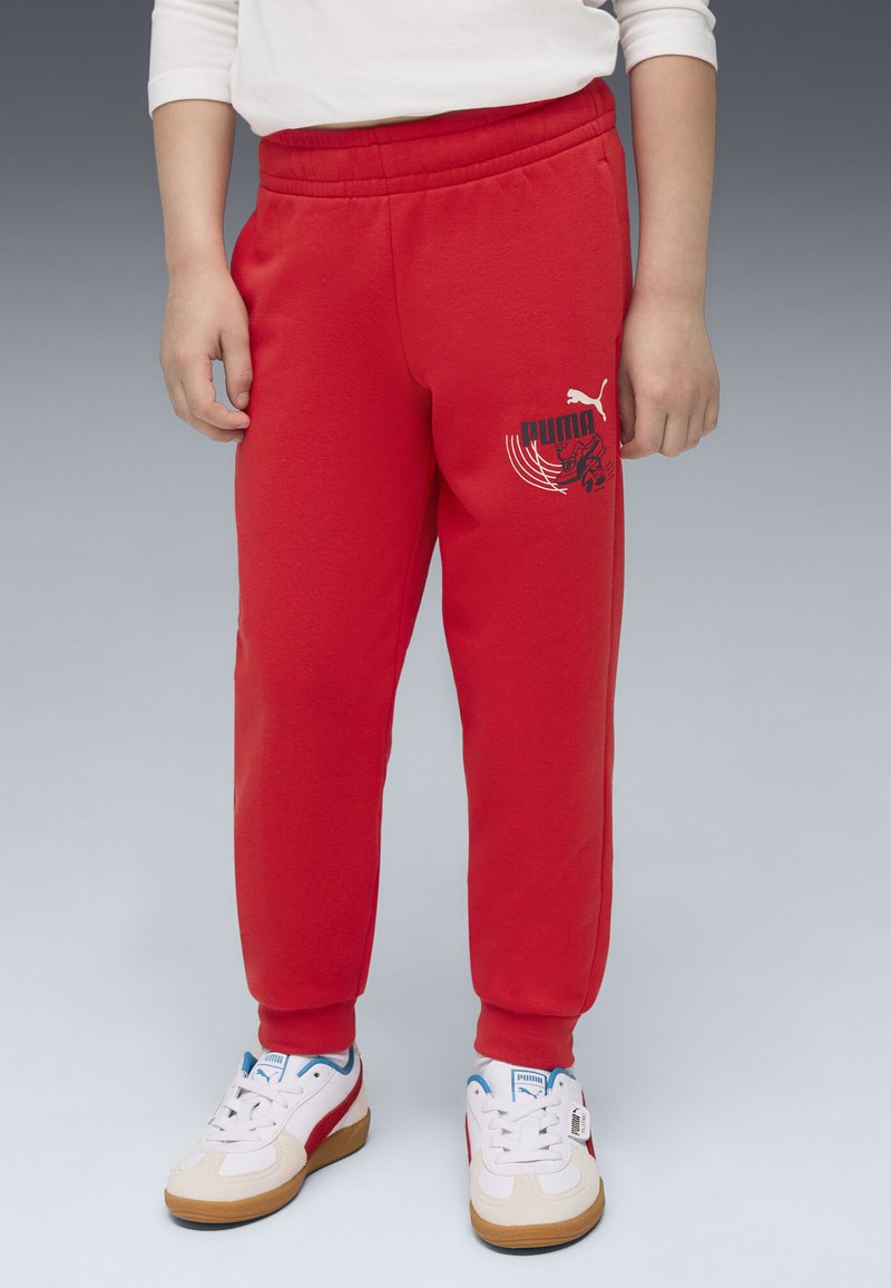Pantaloni da jogging rossi realizzati in tessuto morbido con vita elastica. Presentano un logo grafico nero sulla coscia e polsini a costine. Indossati con sneakers bianche.