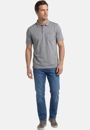 Mann steht, trägt ein graues Polohemd mit kurzen Ärmeln, blaue Jeans und graue Turnschuhe, schaut leicht zur Seite.