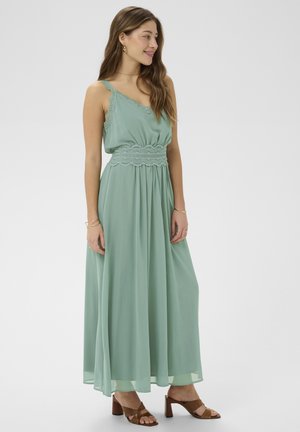 Kobieta nosząca jasnozieloną, bezrękawną sukienkę maxi z koronkowymi detalami oraz brązowe sandały na obcasie, stojąca bokiem i uśmiechająca się.