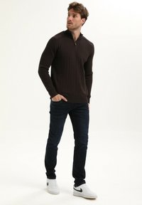 Bruine geribbelde pullover met een half-rits kraag, gecombineerd met donkere jeans en witte sneakers. De pullover heeft lange mouwen en een slim-fit ontwerp.