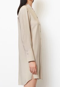 Beiges Langarmhemdkleid aus weichem Stoff, mit einer durchgehenden Knopfleiste, seitlichen Schlitzen und umgeschlagenen Ärmeln mit silbernen Knöpfen.