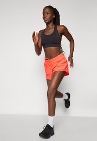 Soutien-gorge de sport noir, shorts néon orange vif avec poches latérales, chaussettes blanches, chaussures de sport noires, modèle féminin courant dans un cadre intérieur.