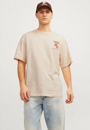 T-shirt en coton beige à manches courtes, avec un imprimé graphique marron sur la poitrine gauche. Un jean en denim bleu clair est porté.